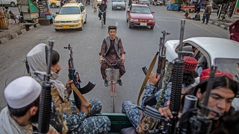 Ein Afghane fährt auf einem Fahrrad, während junge Kämpfer der Taliban auf der Ladefläche eines Pick-Ups in den Straßen von Kabul in Afghanistan patrouillieren. (Foto: picture-alliance / Reportdienste, picture alliance/dpa | Oliver Weiken)