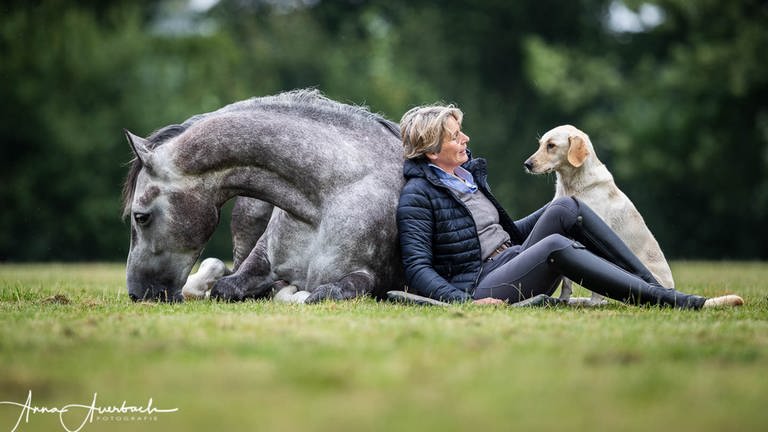 Eine Frau lehnt mit ihrem Rücken an einem Pferd, das es sich auf einer Wiese gemütlich gemacht hat - ein Hund sitzt neben ihr. Das Bild zeigt Anne Krüger-Degener: Sie hat sich auf die Kommunikation mit Tieren spezialisiert - in ihrer Tierschule. Sie ist überzeugt: Dialog bringt eine gute Beziehung zwischen Mensch und Tier. (Foto: Anna Auerbach)