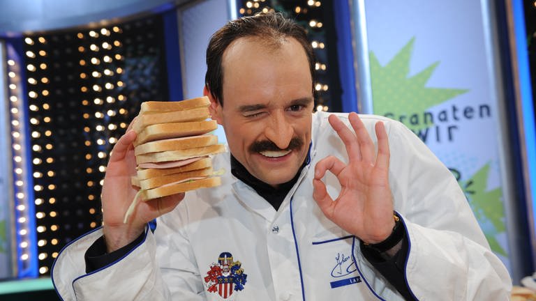 Max Giermann trägt die weiße Kleidung eines Kochs. In der rechten Hand hält er ein überdimensioniertes Sandwich mit Schinken und Salat.  (Foto: picture-alliance / Reportdienste, Jörg Carstensen)