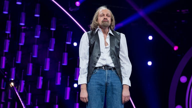 Max Giermann steht auf einer Bühne in Jeans, einem Hemd und einer Lederweste. Er lässt seine Schultern hängen und zieht die Mundwinkel nach unten. (Foto: dpa Bildfunk, Picture Alliance)