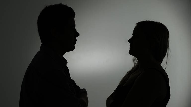 Ein als Silhouette abgebildeter Mann und eine Frau stehen sich mit verschränkten Armen gegenüber. 