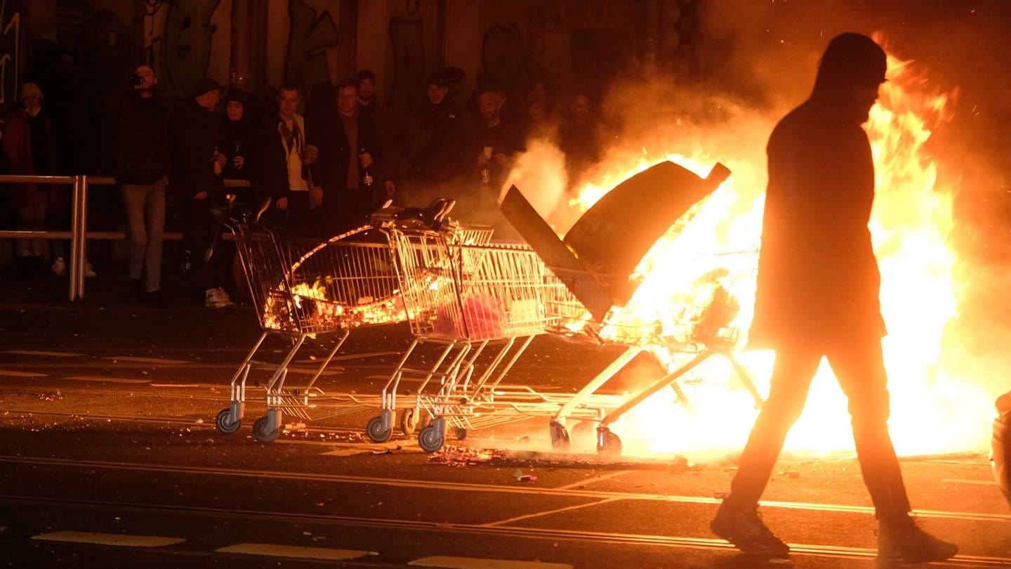 Ein Mann geht an Einkaufwagen mit brennenden Gegenständen vorbeit. Im Osten von Berlin ist es in der Neujahrsnacht zu Ausschreitungen gekommen. (Foto: picture-alliance / Reportdienste, picture alliance/dpa/dpa-Zentralbild | Sebastian Willnow)