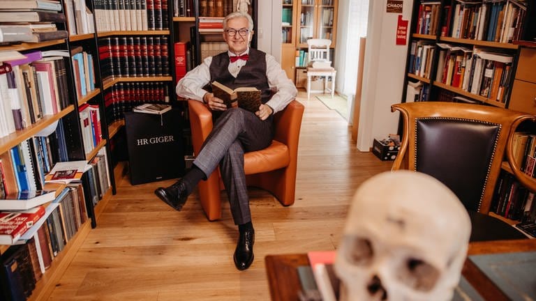 Pathologe Roland Sedivy sitzt vor vollen Bücherregalen in einem Sessel, er hält ein Buch in der Hand und schaut in die Kamera. In der Pathologie arbeitet er an der Diagnose von Krebs und anderen Krankheiten oder blickt in eine Leiche. In SWR1 Leute verrät der Pathologe seine bizarrsten Entdeckungen bei einer Autopsie.
