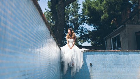 Eine junge Frau im weißen Brautkleid sitzt am Rand eines leeren Schwimmbeckens und sieht mäßig glücklich aus (Foto: IMAGO, IMAGO / Addictive Stock)