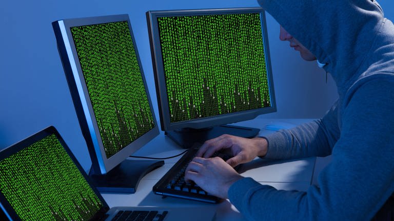 Hacker im Hoodie sitzt vor  drei Monitoren mit Zahlenkolonen (Foto: IMAGO, Panthermedia/Andrey Popov)