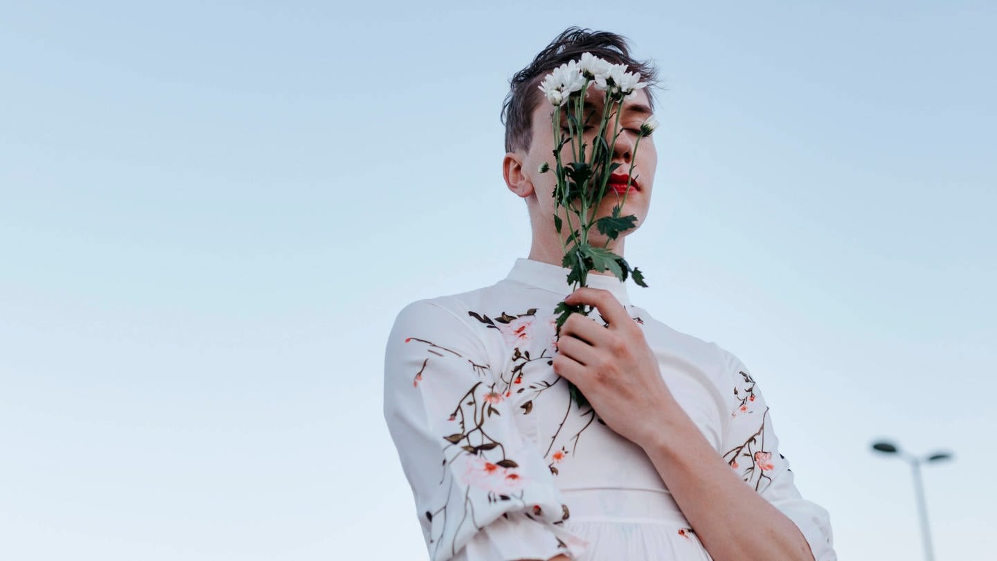 Gender fluid man covering face with bouquet of daisies while standing on street during sunset.