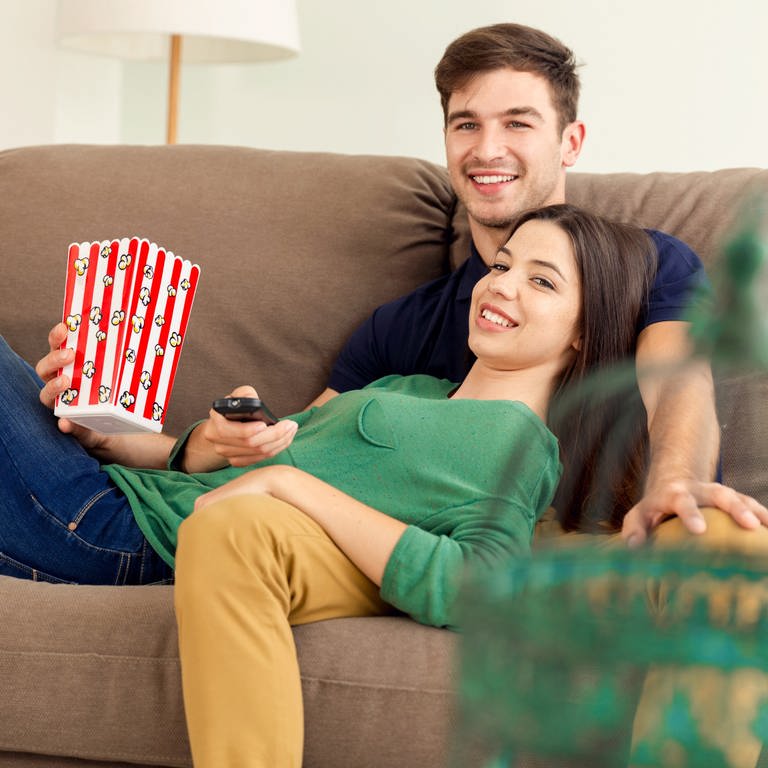 Ein Paar sieht gemeinsam auf der Couch Fernsehen und isst Popcorn (Foto: IMAGO, imago images/ikophotos)