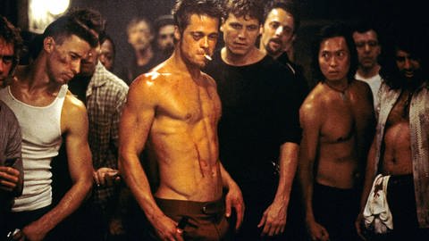 Eine Gruppe Männer steht in einem schlecht beleuchteten Raum im Kreis, in ihrer Mitte Brad Pitt, jung, mit freiem Oberkörper, er blutet am muskulösen bacuh. Er hat eine Zigarette im Mund und ein angeschwollenes Auge. (Foto: IMAGO, IMAGO / Everett Collection)