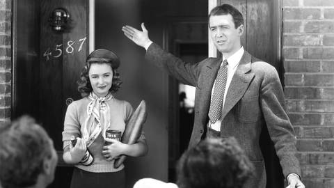 Eine Aufnahme aus einem Film in schwarz-weiß mit James Stewart und Donna Reed vor einem Hauseingang. Stewart gestikuliert im Anzug und zeigt auf eine Austür. (Foto: IMAGO, IMAGO / Everett Collection)
