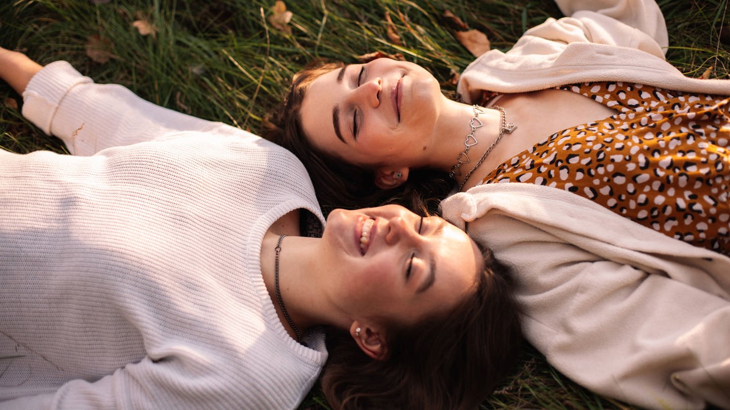 Zwei gückliche Mädchen im Teenageralter lächeln, während sie mit geschlossenen Augen auf dem Gras liegen (Foto: IMAGO, Cavan Images)
