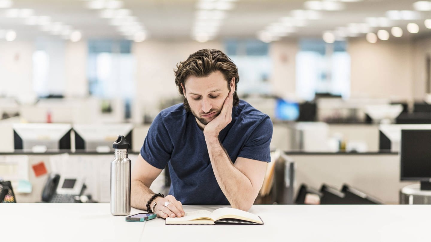 Ein Mann stützt sich auf seinen Ellenbogen und betrachtet ein aufgeschlagenes Buch auf einem Schreibtisch (Foto: IMAGO, Mint Images)