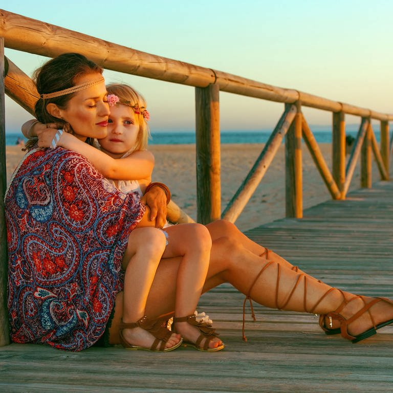 Als Hippie gekleidete Mutter hält Tochter im Arm, am Strand (Foto: IMAGO, agefotostock)