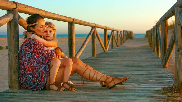 Als Hippie gekleidete Mutter hält Tochter im Arm, am Strand (Foto: IMAGO, agefotostock)