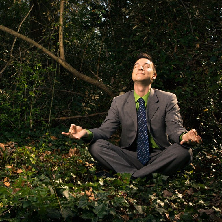 Young businessman sits in a lotus position meditating in the woods with closed eyes and a smile. (Foto: IMAGO, Design Pics)