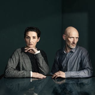 Die Schauspieler*innen Sascha Icks und Tilo Werner sitzen an einer schwarzen, spiegelnden Tischplatte (Foto: NDR)