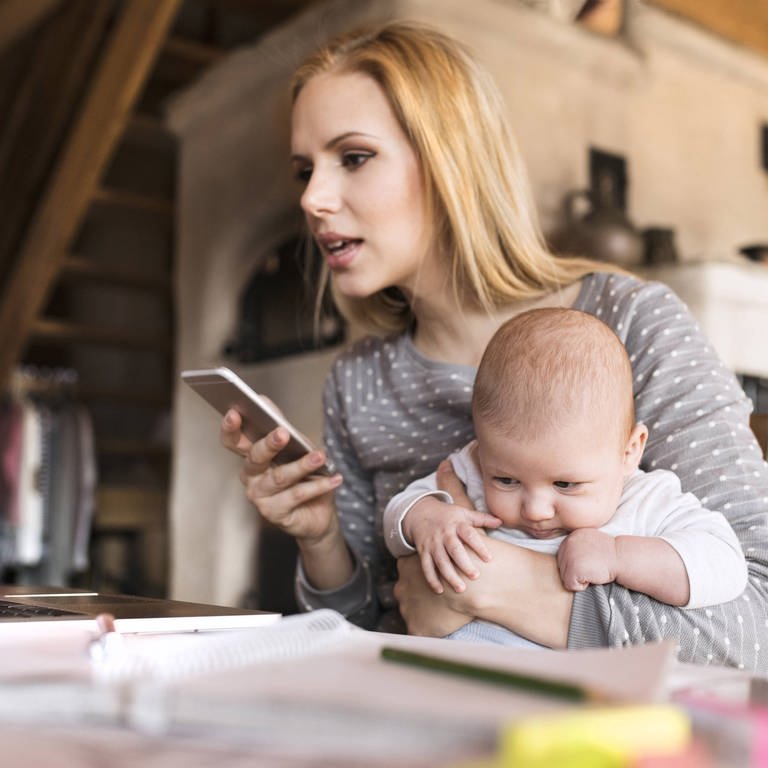 Eine Frau sitzt mit Baby im Arm und Smartphone in der Hand vor einem Notebook (Foto: IMAGO, imago images / Westend61)