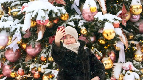 IMAGO  ITAR-TASS (Foto: imago images, Eine warm angezogene Frau mit Mütze und Pelzmantel macht ein Selfie vor einem reich geschmückten Weihnachtsbaum in Russland.)