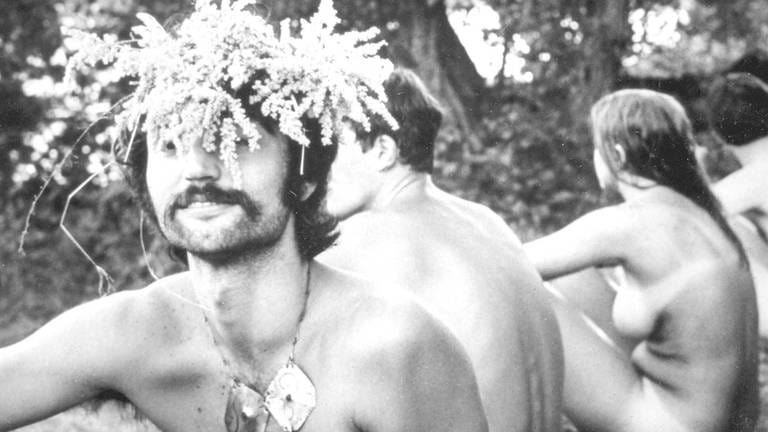 Hippies sitzen nackt im Gras, ein junger Mann trägt einen Blumenkranz auf dem Kopf (Foto: IMAGO, EntertainmentPictures)