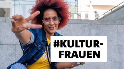 Junge Frau mit einem rot gefärbten Afro sitzt auf einer Treppe und zeigt das V-Zeichen mit der rechten Hand. Links hält sie ein Smartphone. Auf dem Bild ist ein Textblock #Kultufrauen, schwarze Schrift auf weißem Grund. (Foto: IMAGO, Imago / Cavan Images)