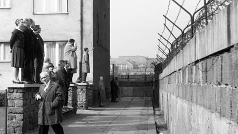 Eine Reisegruppe schaut von einer Vorgartenmauer über die Mauer hinweg vom Westteil in den Ostteil von Berlin, Deutschland 1962: Schwarz-weiß Bild, links stehen die Menschen, Männer und Frauen auf einem kleinen Mäuerchen, rechts befindet sich in mehreren Metern Abstand die Mauer, die oben mit Stacheldraht gesichert ist. (Foto: IMAGO, IMAGO / United Archives)