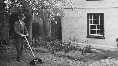 Ein älterer Mann schneidet seinen Rasen mit einem handbetriebenen Rasenmäher in Kent, England, am 7. April 1938: Ein bühender Baum im Hintergrund, der Mann trägt einen Bart, einen Anzug mit Hut und schiebt den Rasenmäher vor sich. Hinter ihm ist noch ein Haus zu sehen. Das Bild ist schwraz-weiß. (Foto: IMAGO, IMAGO / United Archives International)