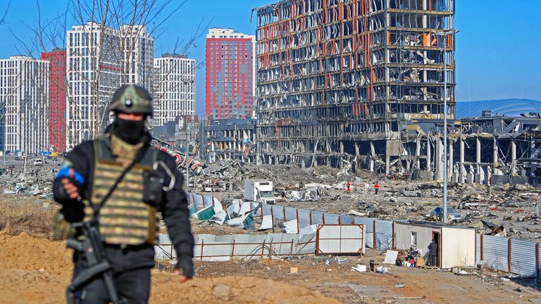 Ein unscharfer Soldat vor einer zerstörten Skyline. Moderne Glas- und Betonbauten sind großflächig zerstört. Davor laufen Rettungskräfte herum. (Foto: IMAGO, IMAGO / NurPhoto)