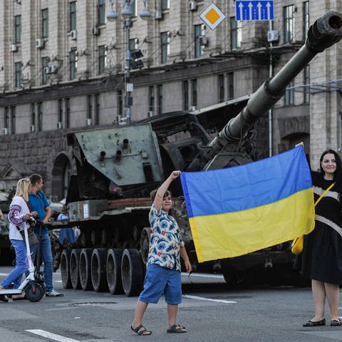 Eine Frau und ein junge machen Fotos mit der ukrainischen Flagge vor einem zerstörten russichen Panzer in Kiew (Foto: IMAGO, IMAGO / ZUMA Wire)