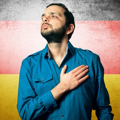 Ein junger Mann - mit rechter Hand auf der linken Brust - steht vor einer Deutschland-Fahne (Foto: IMAGO, PantherMedia / George Mdivanian)