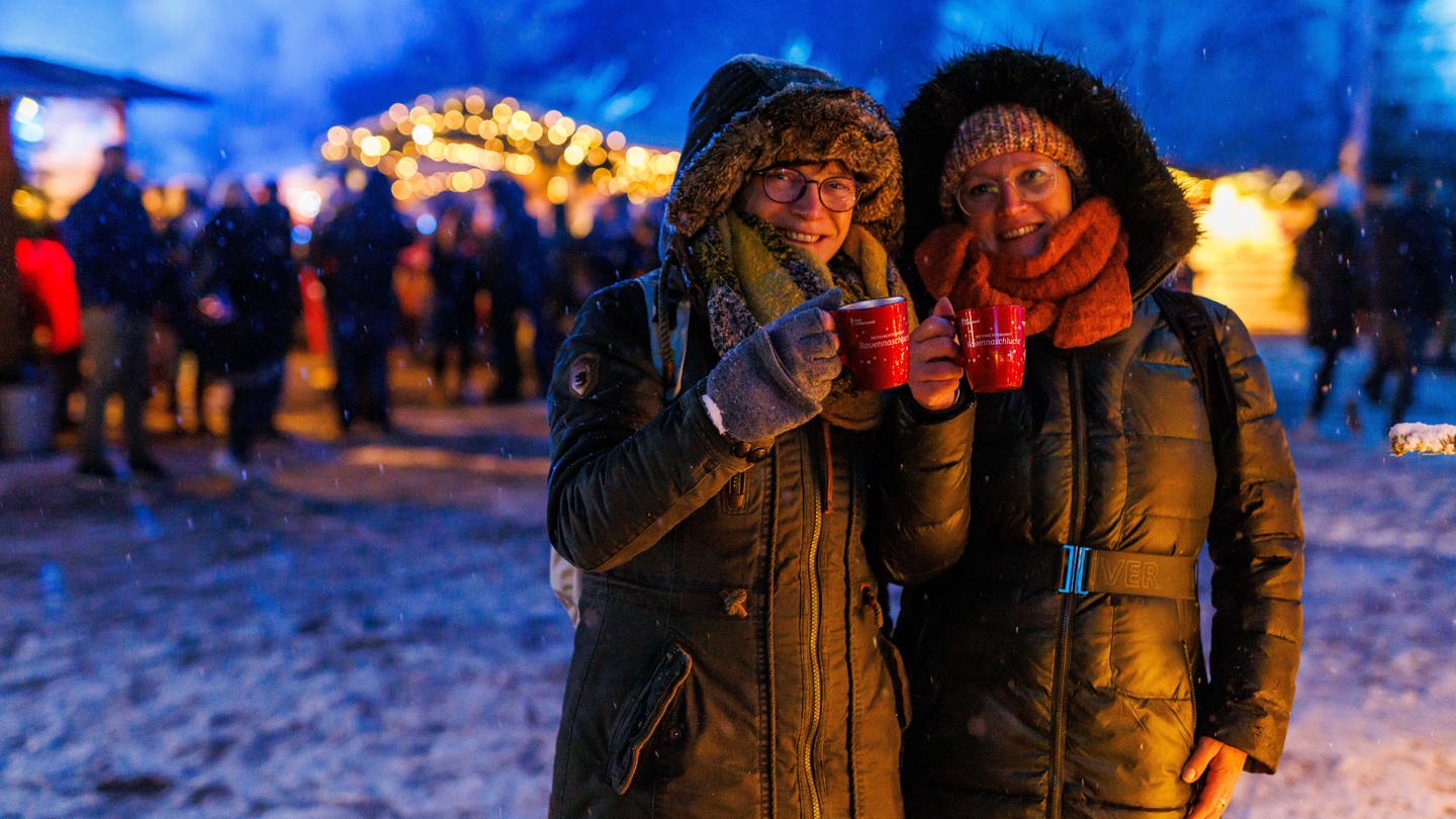 Zwei Frauen stehen mit zwei Tassen Glühwein in der Hand auf dem Weihnachtsmarkt.