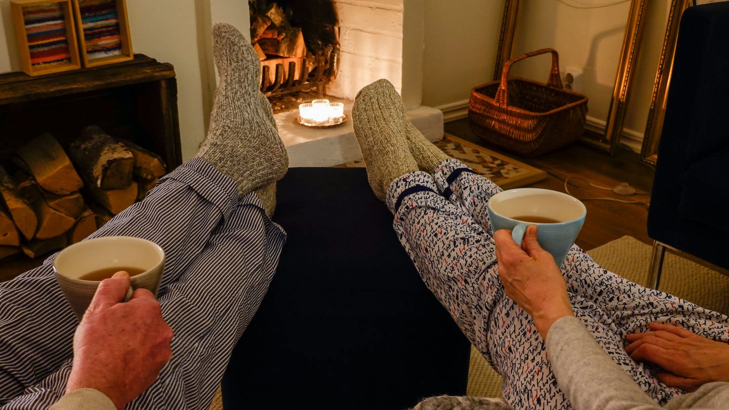 A couple sit together drinking tea with wool socks in front of fireplace Symbolfoto