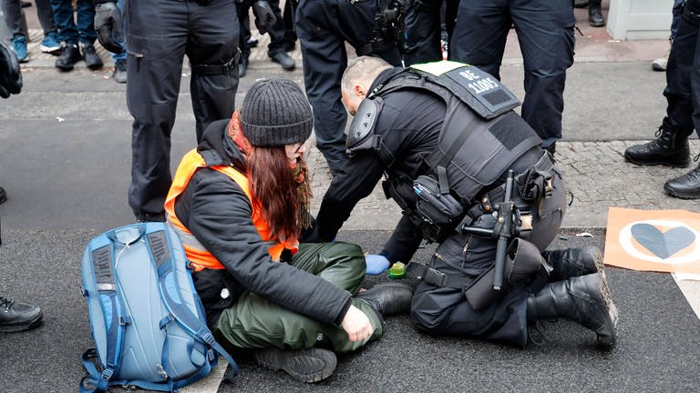 Eine Frau klebt sich auf der Straße fest und ein Polizist versucht sie zu entfernen (Foto: IMAGO, IMAGO/Future Image)