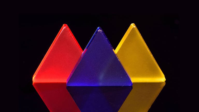 Dreiecke aus rotem, blauem und gelbem Acrylglas auf spiegeldem schwarzen Untergrund. (Foto: IMAGO, CHROMORANGE)