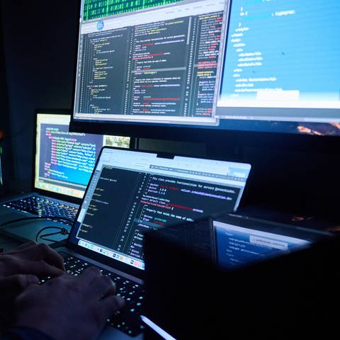 Illustration: Fiktive html-Seiten und Hacker-Programme sind auf Bildschirmen zu sehen, während ein Mann seinen Hände auf der Tastatur hat. (Foto: picture-alliance / Reportdienste, picture alliance/dpa | Annette Riedl)