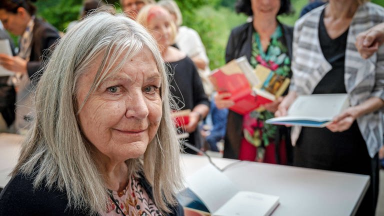 Die Autorin Helga Schubert: Links im Bild eine ältere grauhaarige weiße Frau mit strähnigem schulterlangem grauem Haar. Sie lächelt in die Kamera, im Hintergrund unscharf stehen Menschen an einem Tisch an, um sich Bücher signieren zu lassen. (Foto: IMAGO, IMAGO / Eberhard Thonfeld)