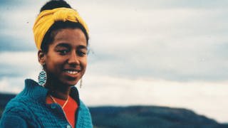 Die Schriftstellerin May Ayim: Eine junge schwarze Frau lächelt in die Kamera, ihre Haare sind nach oben gebunden, sie trägt ein gelbes Haarband, große Ohrhänger, einen blauen Strickpullover. Im V-Ausschnitt des Strickpullover sieht man ein rotes T-Shirt und eine goldene Kette. Im Hintergrund ist hügelig-waldige Natur unscharf zu erkennen, der Himmel ist gräulich-bewölkt. (Foto: Dagmar Schultz / FFBIZ)