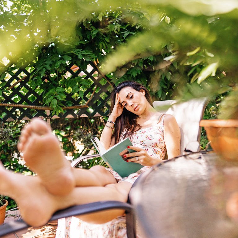 Eine junge, schwangere, schwarzhaarige Frau liegt auf einem Liegestuhl und liest in einem grünen Buch. Sie trägt ein beiges langes Kleid mit Blumenmuster und stützt ihren Kopf in der rechten Hand ab. Sie ist von vielen grünen Pflanzen umgeben. Ihre Fußsohlen sind unscharf im Vordergrund zu sehen. (Foto: IMAGO, IMAGO / Addictive Stock)