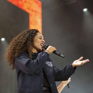 Die Sängerin Joy Denalane während eines Konzerts, sie hält ein Mikrofon in der Hand und singt, sie ist eine hellhäutige schwarze Frau mit langen, braunen gelockten Haaren, sie trägt eine dunkelblaue Jacke mit weißem Logo. (Foto: imago images, IMAGO / Marcel Lorenz)