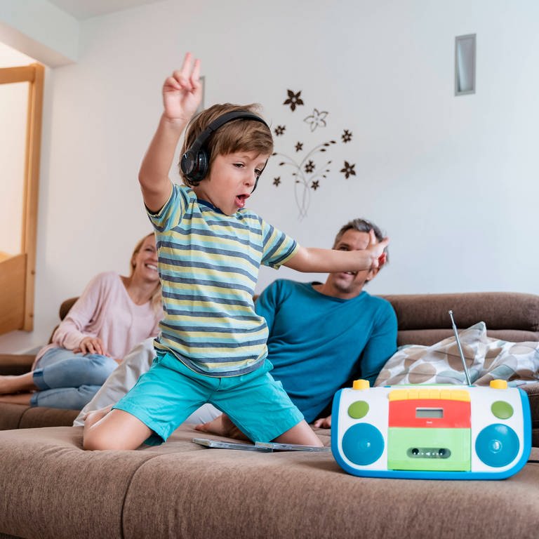 Junge hört mit Kopfhörern Musik während seine Eltern im Hintergrund auf dem Sofa sitzen (Foto: IMAGO, IMAGO / Westend61)