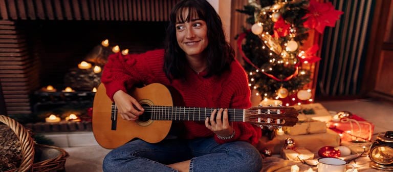 Young woman playing guitar while sitting at home during Christmas (Foto: IMAGO, IMAGO / Westend61)