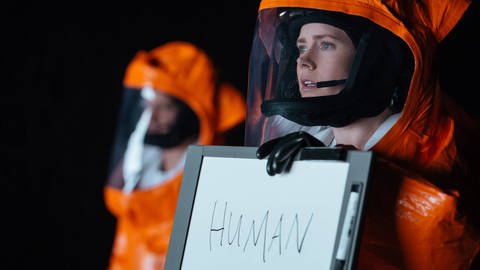 Amy Addams in einem Schutzanzug, sie hält ein Schild mit der Aufschrift "Human" in der Hand, ein Still aus "Arrival" (2016) (Foto: IMAGO, Imago / Entertainment Pictures / ZUMAPRESS)