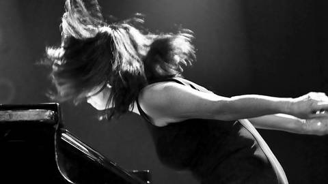 Frau am Flügel, die vom Klavierhocker springt und die Hände nach hinten reißt; die Haare fallen wild ins Gesicht (Foto: Pressestelle, Ziga Koritnik)