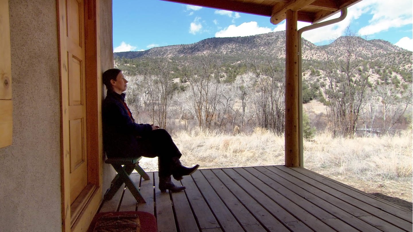 Auf dem Foto sieht man Meredith Monk von der Seite  sitzend auf der Terasse eines Hauses. Im Hintergrund sind Berge zu sehen.