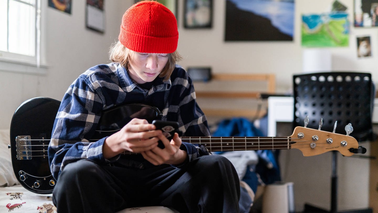 Junge mit rotem Beany hält eine Gitarre und das Smartphone in der Hand (Foto: IMAGO, IMAGO / Cavan Images)