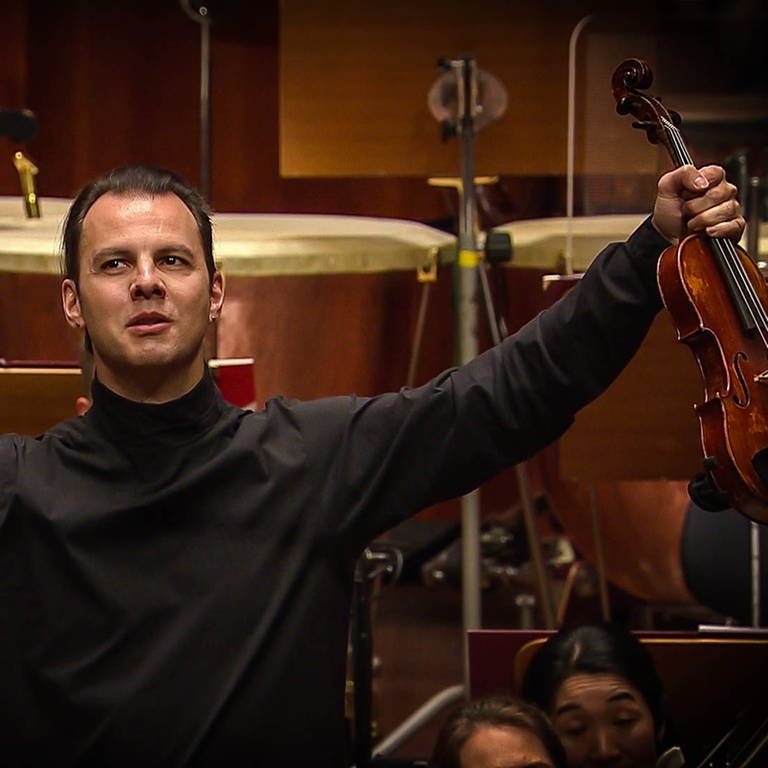 Teodor Currentzis mit ausgestreckten Armen,  hält Geige und Bogen in den Händen (Foto: SWR)