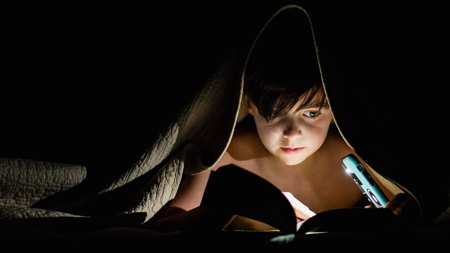 Ein Kind liegt im Bett und liest unter der Decke ein Buch bei Nacht (Foto: IMAGO, IMAGO / Cavan Images)
