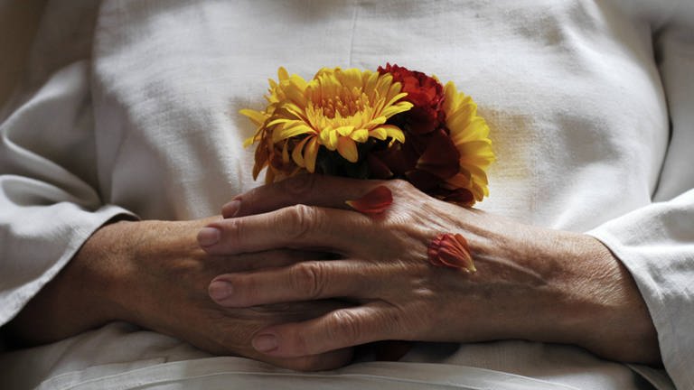Hände einer Toten mit Blumen: Die meisten Menschen möchten zu Hause sterben; seit 2008 haben sie das Recht, dort palliativmedizinisch begleitet zu werden. (Foto: IMAGO, imago images / photothek)