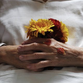 Hände einer Toten mit Blumen: Die meisten Menschen möchten zu Hause sterben; seit 2008 haben sie das Recht, dort palliativmedizinisch begleitet zu werden. (Foto: imago images, imago images / photothek)