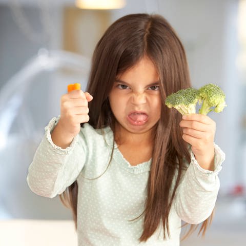 Kleines Mädchen schneidet wilde Grimasse und hält Brokkoli und Möhre von sich: Wie etwas schmeckt, legen nicht nur Zunge und Nase fest. Auch unsere Erfahrungen bestimmen darüber, vor allem aber die Kultur, in der wir leben.  (Foto: IMAGO, IMAGO / Shotshop)