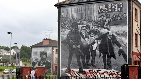 Häuser mit Wandgemälden in Derry, die an den "Bloody Sunday", den "Blutsonntag" am 30. Januar 1972, erinnern (Foto: picture-alliance / Reportdienste, Häuser mit Wandgemälden in Derry, die an den Bloody Sunday, den Blutsonntag am 30. Januar 1972, erinnern. Die Szene im Vordergrund zeigt Father Daly mit einem weißen blutbefleckten Taschentuch winkend, der die kleine Gruppe mit dem sterbenden Jackie Duddy anführt.)