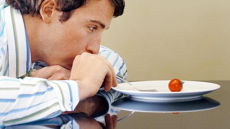 Mann sitzt über einen Teller gebeugt und stochert mit seiner Gabel auf einem Teller mit nur einer kleinen Cocktail-Tomate herum. (Foto: Getty Images, Thinkstock -)